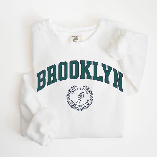 A Comfort Colors lightweight cotton crewneck sweatshirt with "Brooklyn Track & Field" printed in vintage retro style, evoking an old money aesthetic.