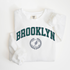 A Comfort Colors lightweight cotton crewneck sweatshirt with "Brooklyn Track & Field" printed in vintage retro style, evoking an old money aesthetic.