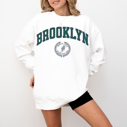 A Comfort Colors lightweight cotton crewneck sweatshirt with "Brooklyn Track & Field" printed in vintage retro style, evoking an old money aesthetic.