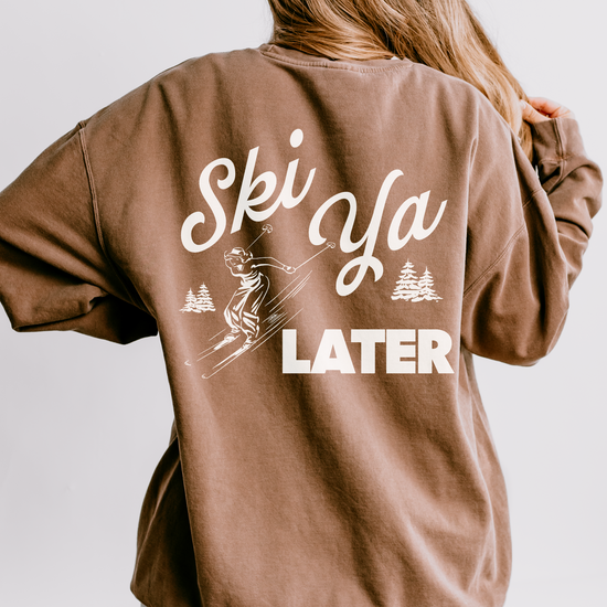 Model wearing a lightweight cotton crewneck sweatshirt with a 'Ski Ya Later' design. The back of the sweatshirt features a vintage boho illustration of a woman skier, adding retro charm to the relaxed, casual fit.