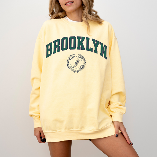 A Comfort Colors lightweight cotton crewneck sweatshirt with "Brooklyn Track & Field" printed in vintage retro style, evoking an old money aesthetic.