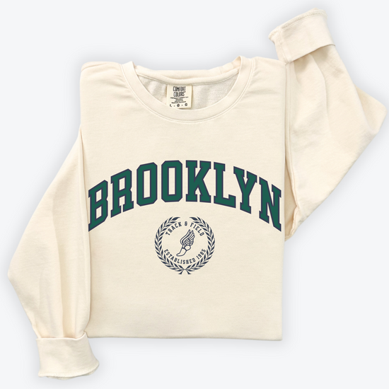 A Comfort Colors lightweight cotton crewneck sweatshirt with "Brooklyn Track & Field" printed in vintage retro style, evoking an old money aesthetic.