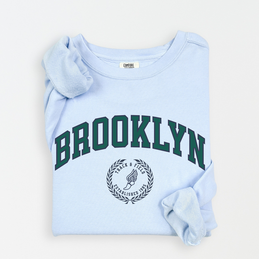 A Comfort Colors lightweight cotton crewneck sweatshirt with "Brooklyn Track & Field" printed in vintage retro style, evoking an old money aesthetic.