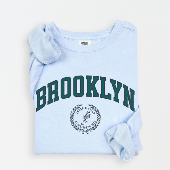 A Comfort Colors lightweight cotton crewneck sweatshirt with "Brooklyn Track & Field" printed in vintage retro style, evoking an old money aesthetic.